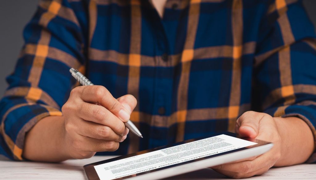 Business professional in plaid shirt using a stylus to sign and annotate a digital document on a tablet, performing detailed contract analysis for review and approval.