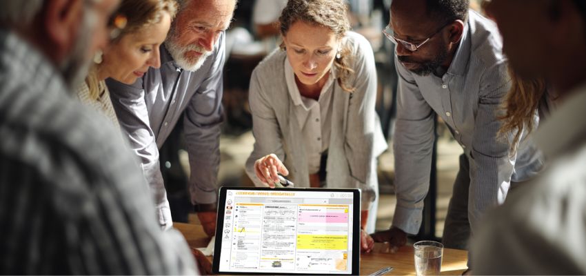 Group of diverse colleagues gathered around a table collaboratively pointing at and discussing a highlighted contract summary displayed on a tablet during a meeting.