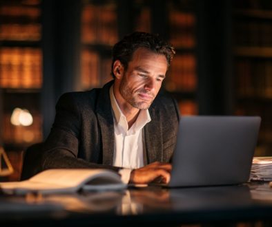 Professional lawyer in a gray suit focused on his laptop screen late at night in a law library, surrounded by stacks of legal documents and a judge’s gavel, illustrating the integration of legal AI into modern legal practice and case preparation